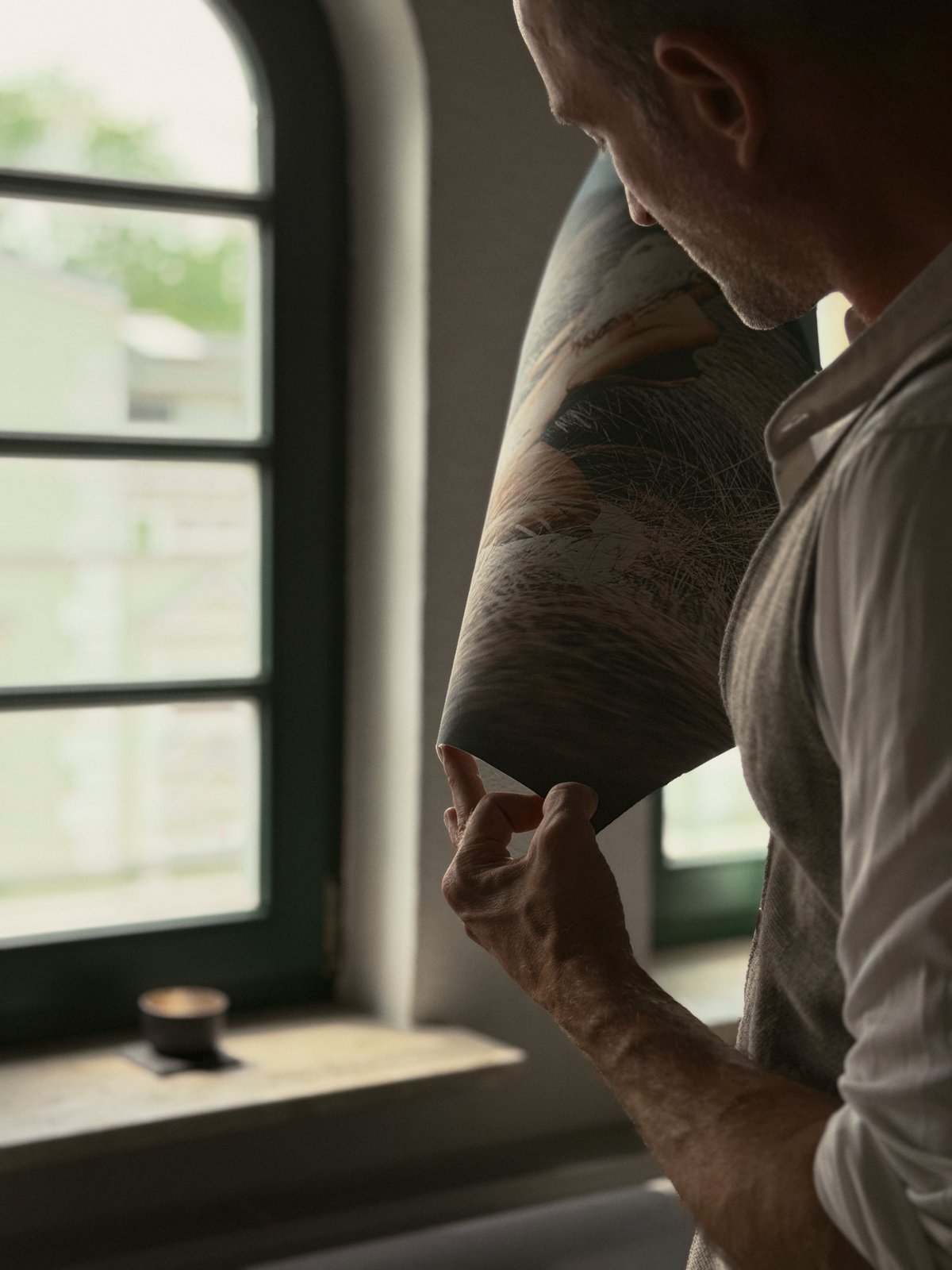 Eine Hand haelt einen aufgerollten Fine-Art-Druck gegen das Fensterlicht, die Papieroberflaeche ist im Streiflicht sichtbar.
